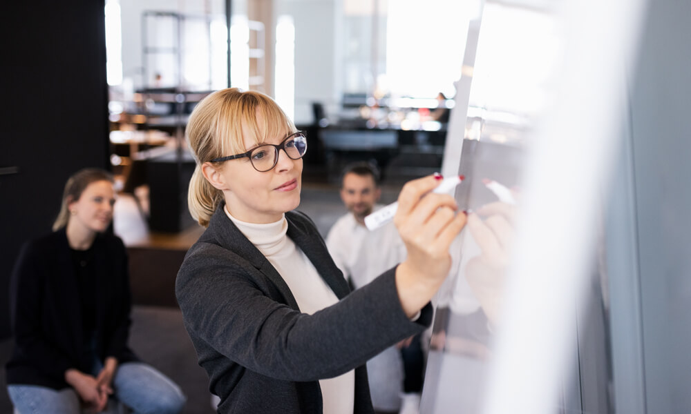 A female team leader writing on a whiteboard while the team members brainstorm